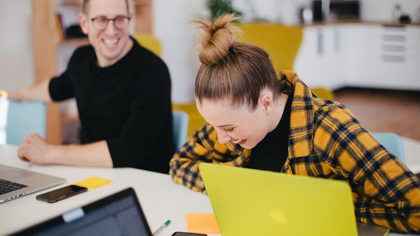 Two people with laptops sitting around a meeting room table and laughing