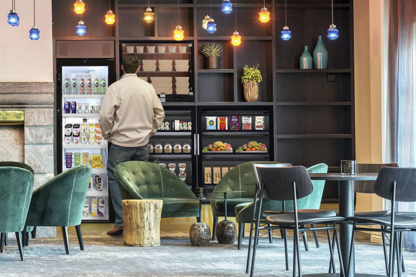 Person browsing self-service refrigerators in a modern café with green velvet chairs and blue and yellow pendant lights.